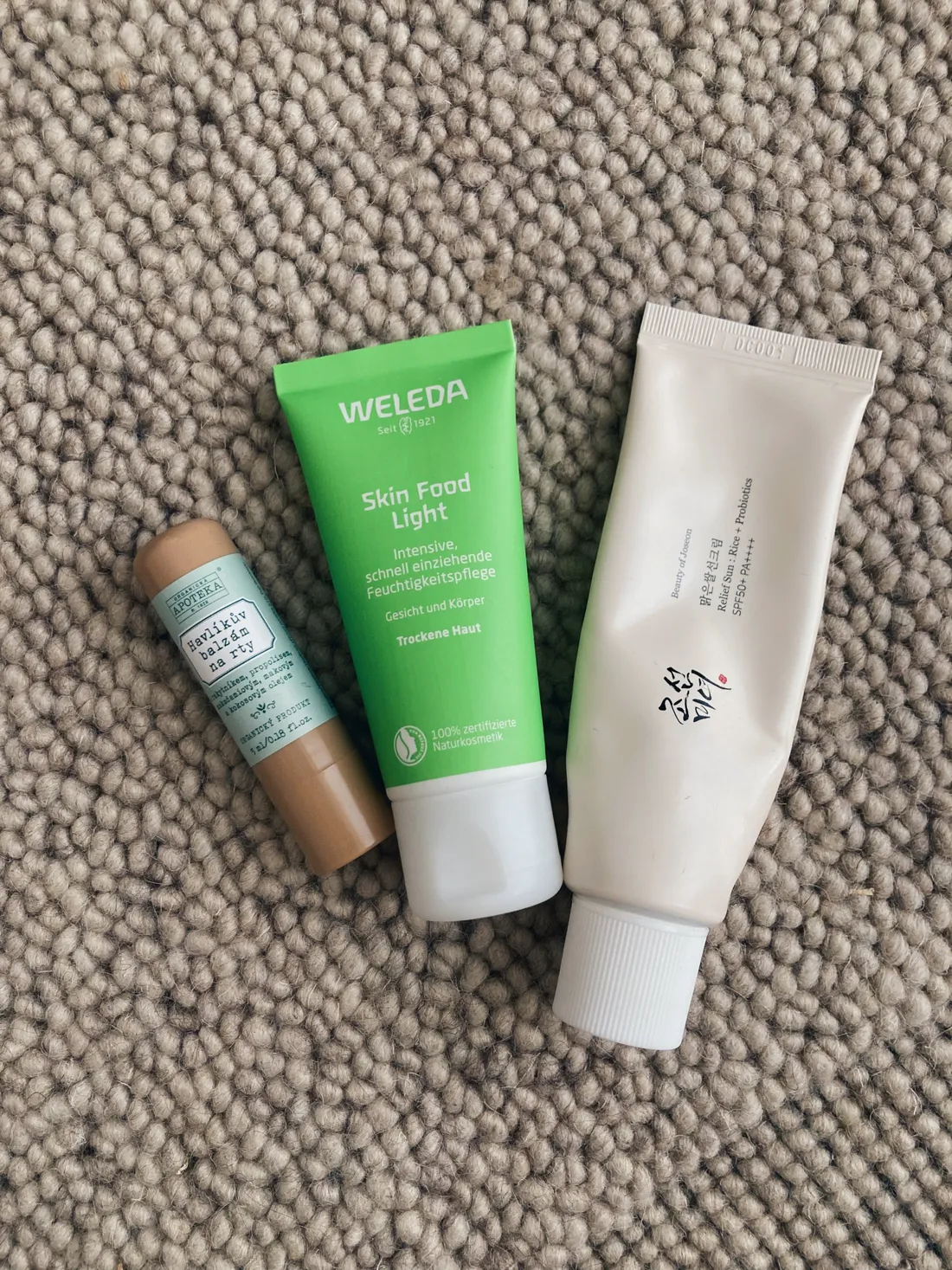 Tři kosmetické produkty Weleda, Havlík a Beauty of Joseon na světle šedém koberci.