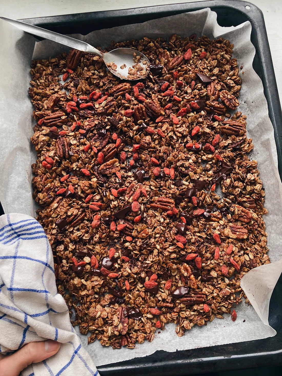 Domácí granola s pekanovými ořechy, goji a čokoládou na plechu s lžící shora.