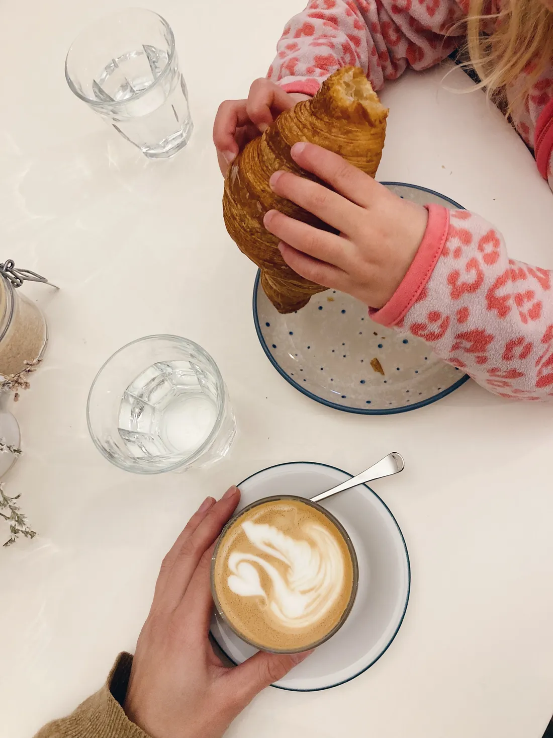 Latte s latte art labutě a croissant držený dítětem na bílém stole shora.