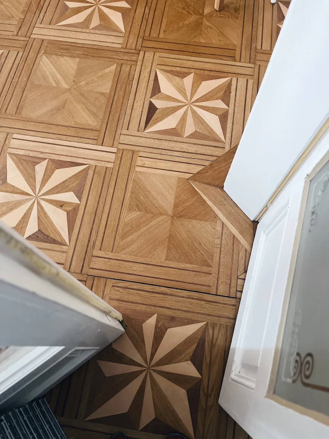 Dřevěná parketová podlaha s geometrickým vzorem osmicípých hvězd a diagonální kresby u bílých dveří.