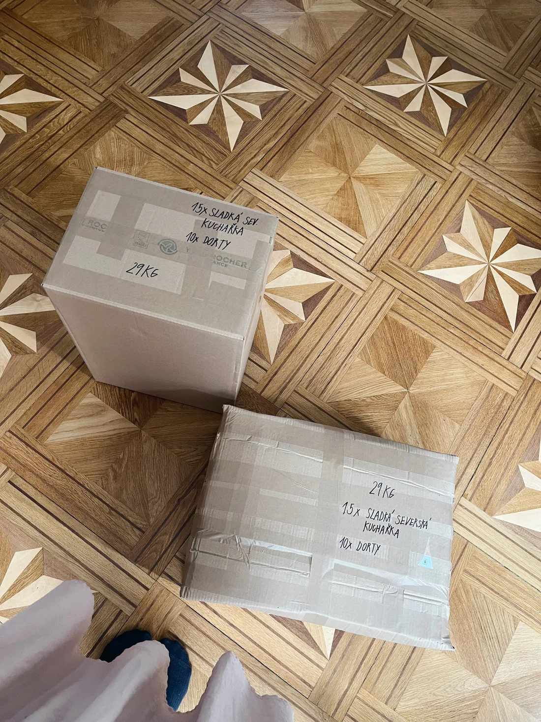 Dvě papírové krabice s ručně psanými nápisy o kuchařkách a váze 29 kg na ozdobné parketové podlaze.