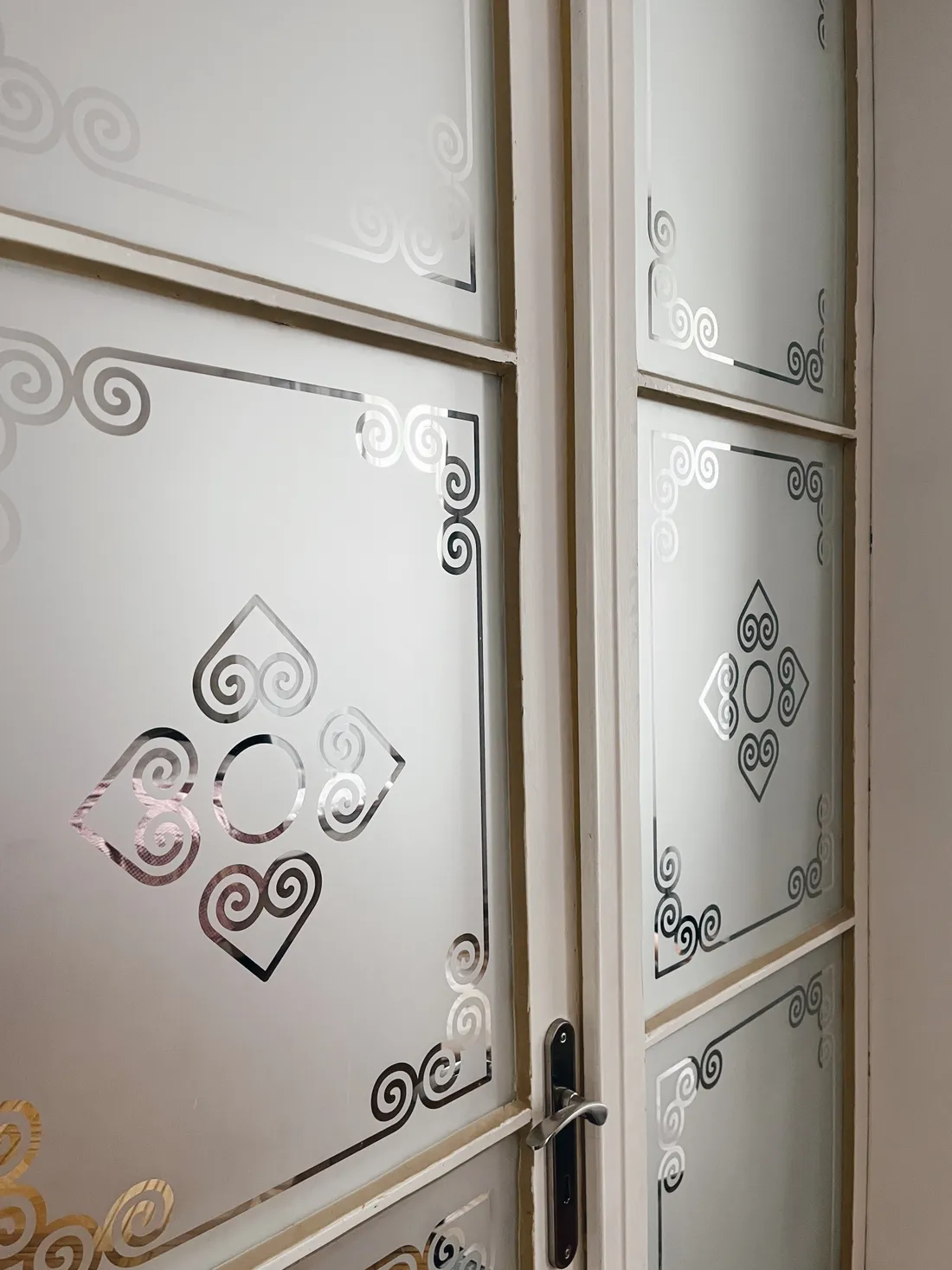 Detail bílých dveří s pískovaným sklem a stříbrnými ornamenty ve tvaru spirál a srdcí.