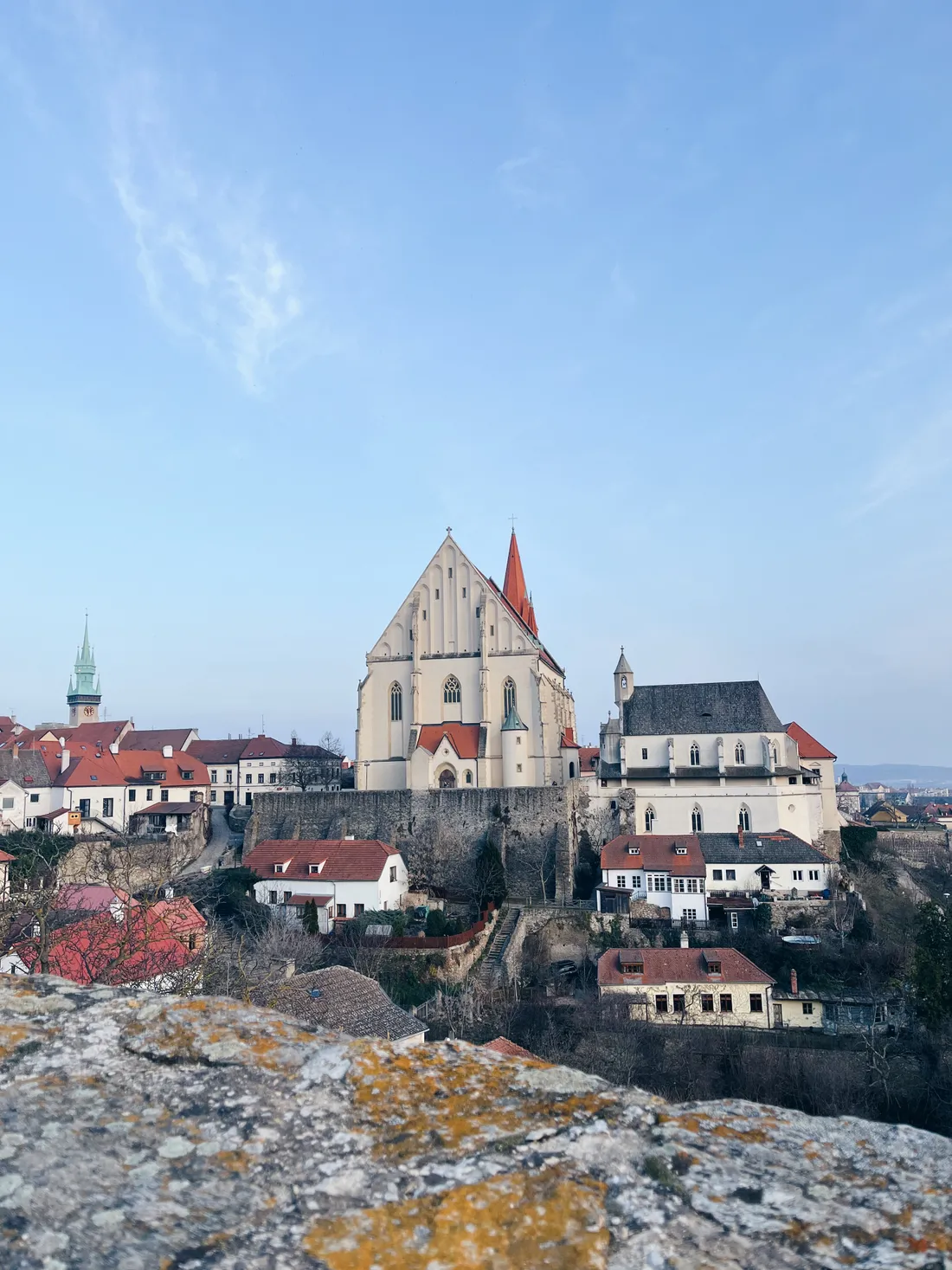 Bílý gotický kostel a kaple na hradbách nad domy s červenými střechami, v popředí kamenná zídka.
