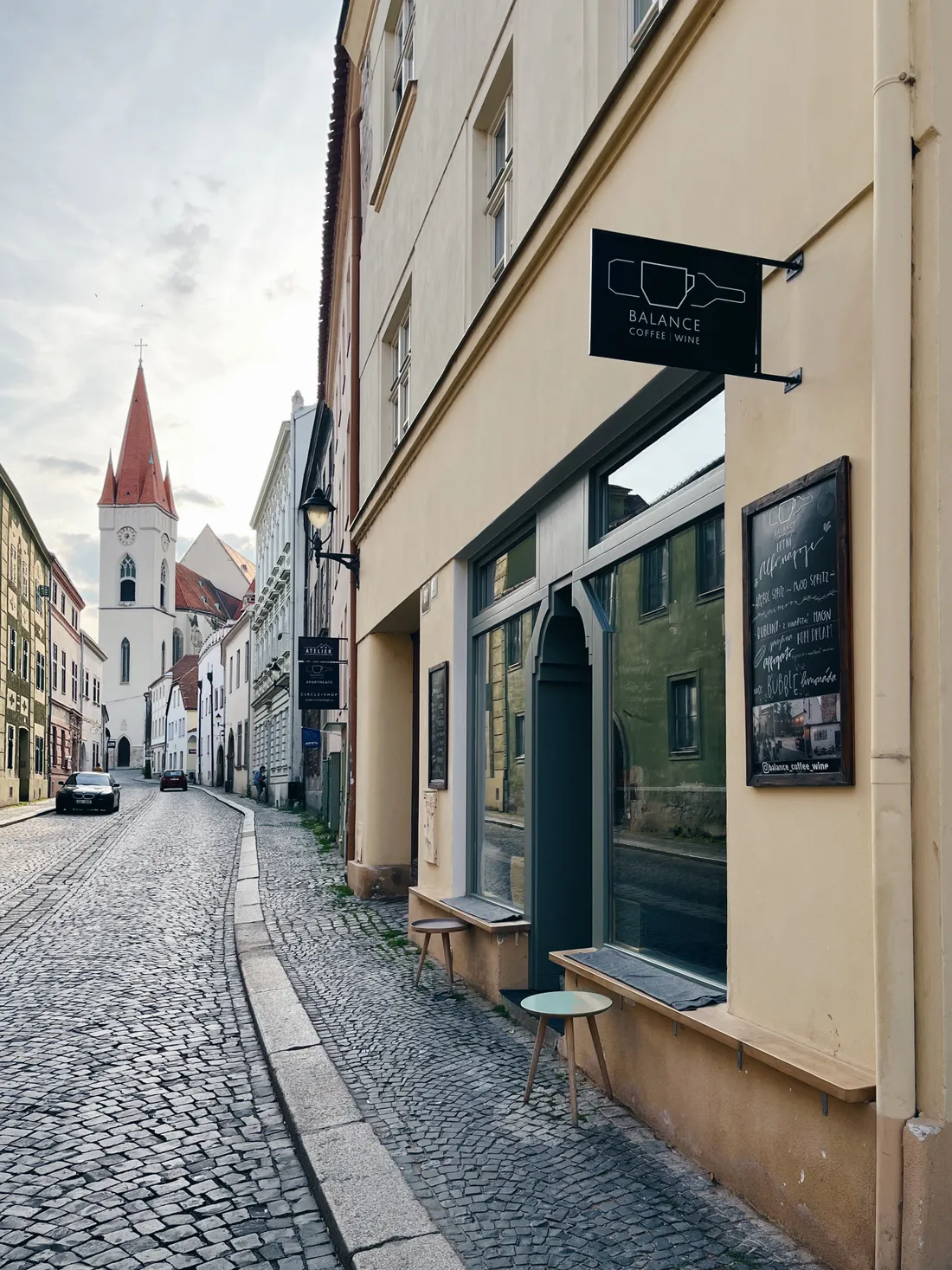 Úzká dlážděná ulice k bílému kostelu, kavárna Balance Coffee Wine se štítkem.