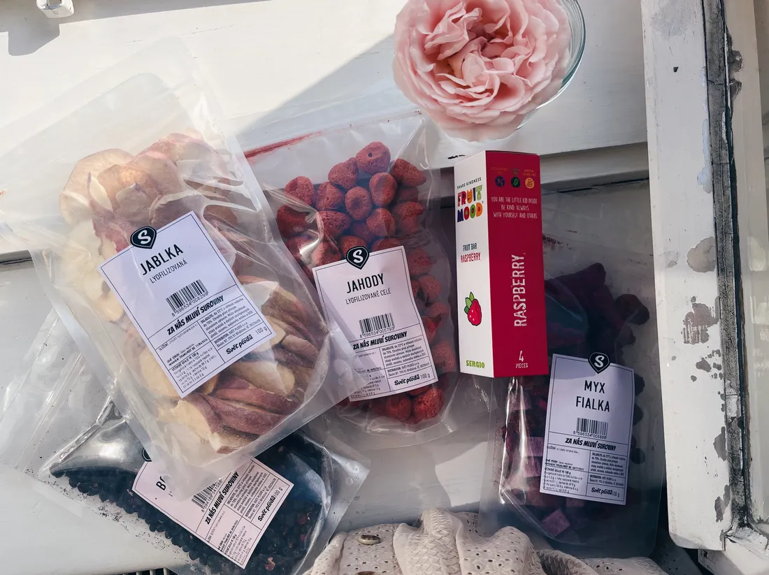 Sáčky lyofilizovaného ovoce a tyčinka Fruit Mood na bílém parapetu vedle světle růžové růže.