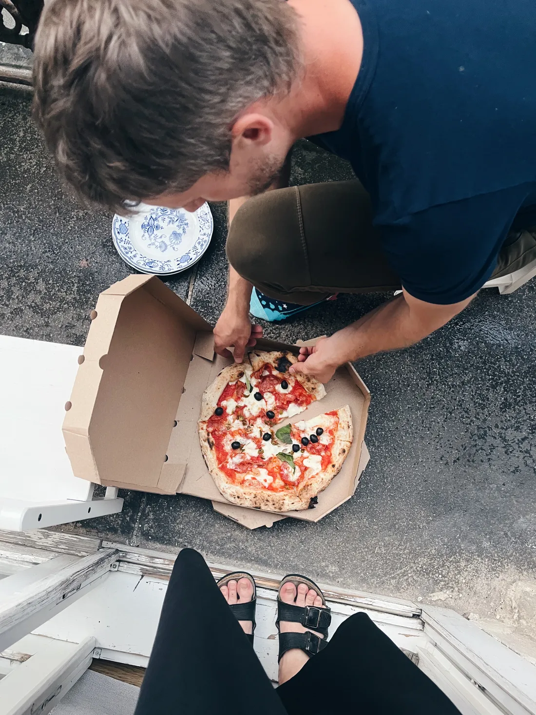 Muž v modrém triku otevírá krabici s pizzou na venkovní dlažbě, v popředí nohy v sandálech.