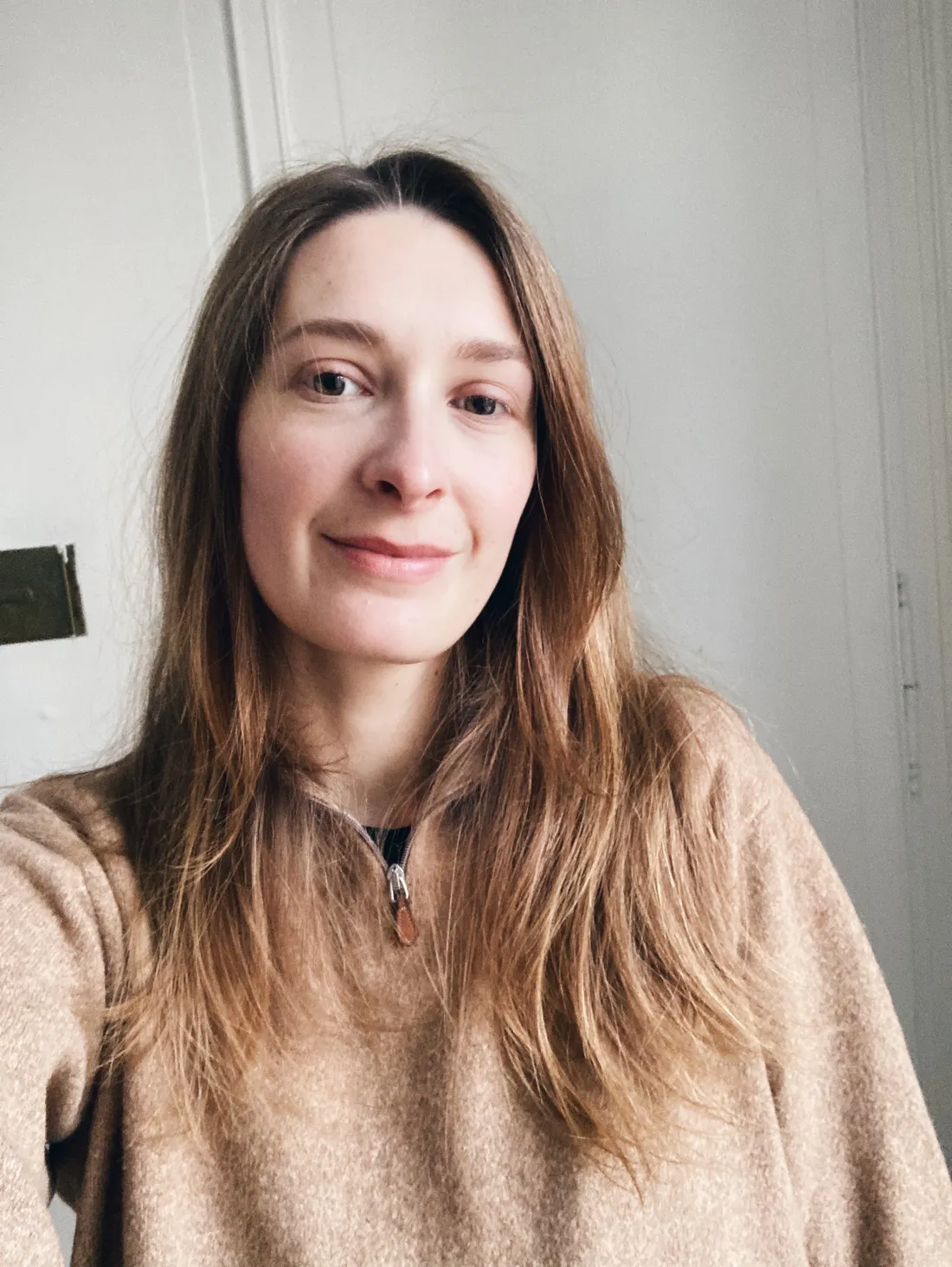 Žena s dlouhými světle hnědými vlasy v béžovém svetru se zipem na světlé stěně.