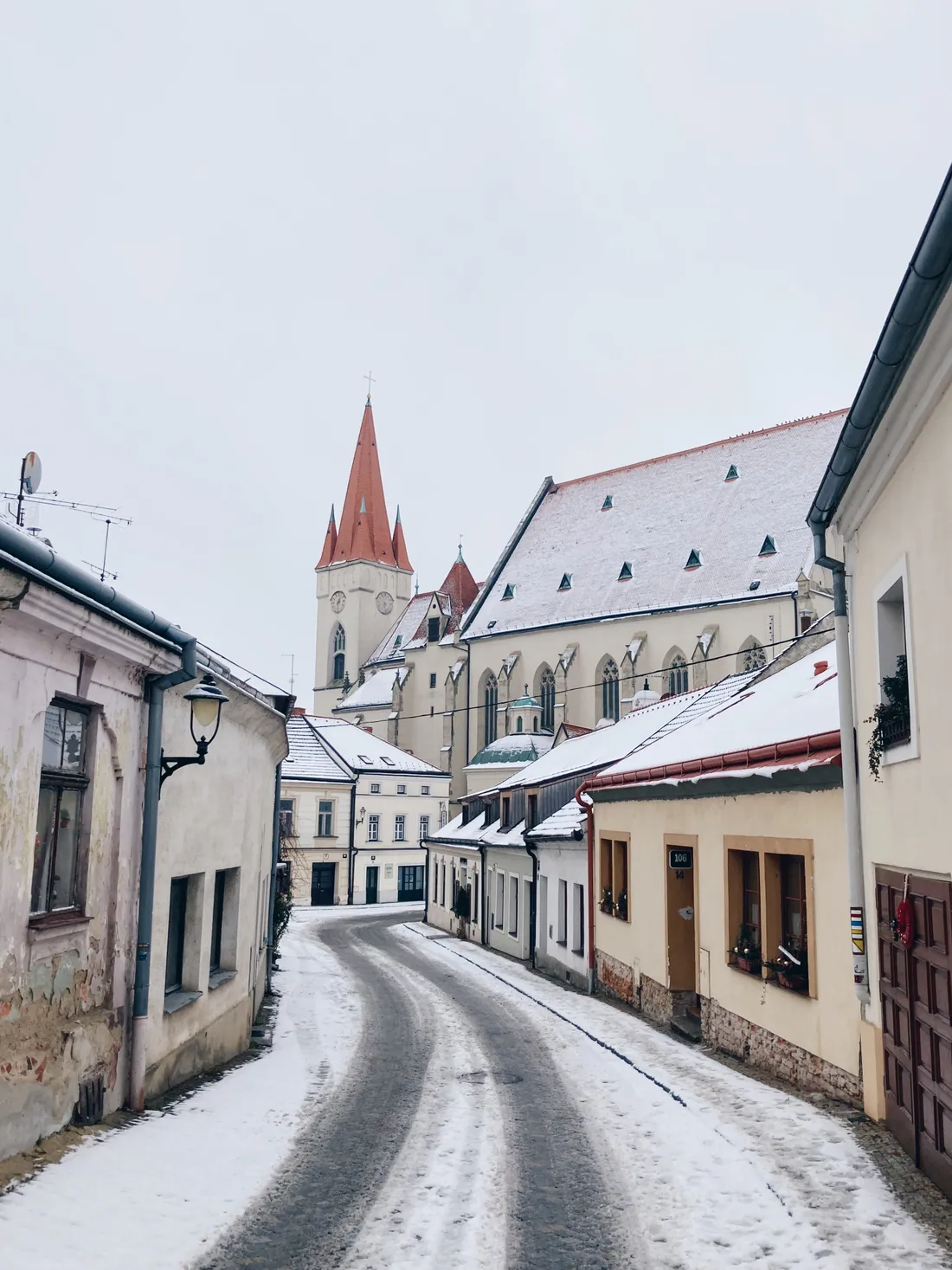 Zasněžená ulička mezi domy vede k gotickému kostelu s vysokou červenou střechou a věží.