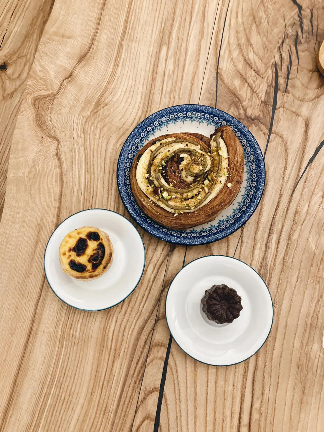 Tři dezerty na talířích: šnek s pistáciemi, pastel de nata a canelé na dřevěném stole s parketami.