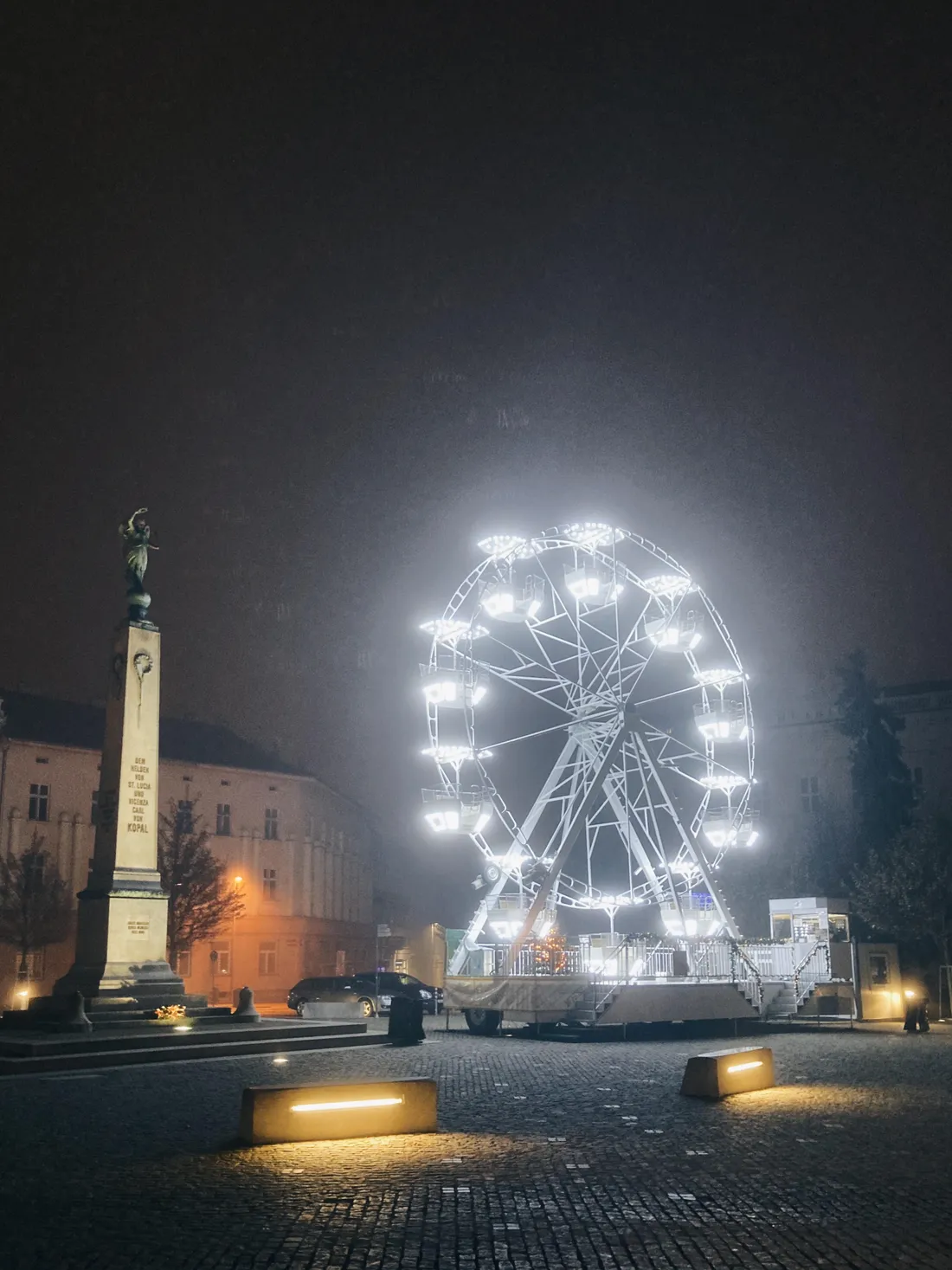 Osvětlené ruské kolo na dlážděném náměstí v noci vedle kamenného památníku v mlze.