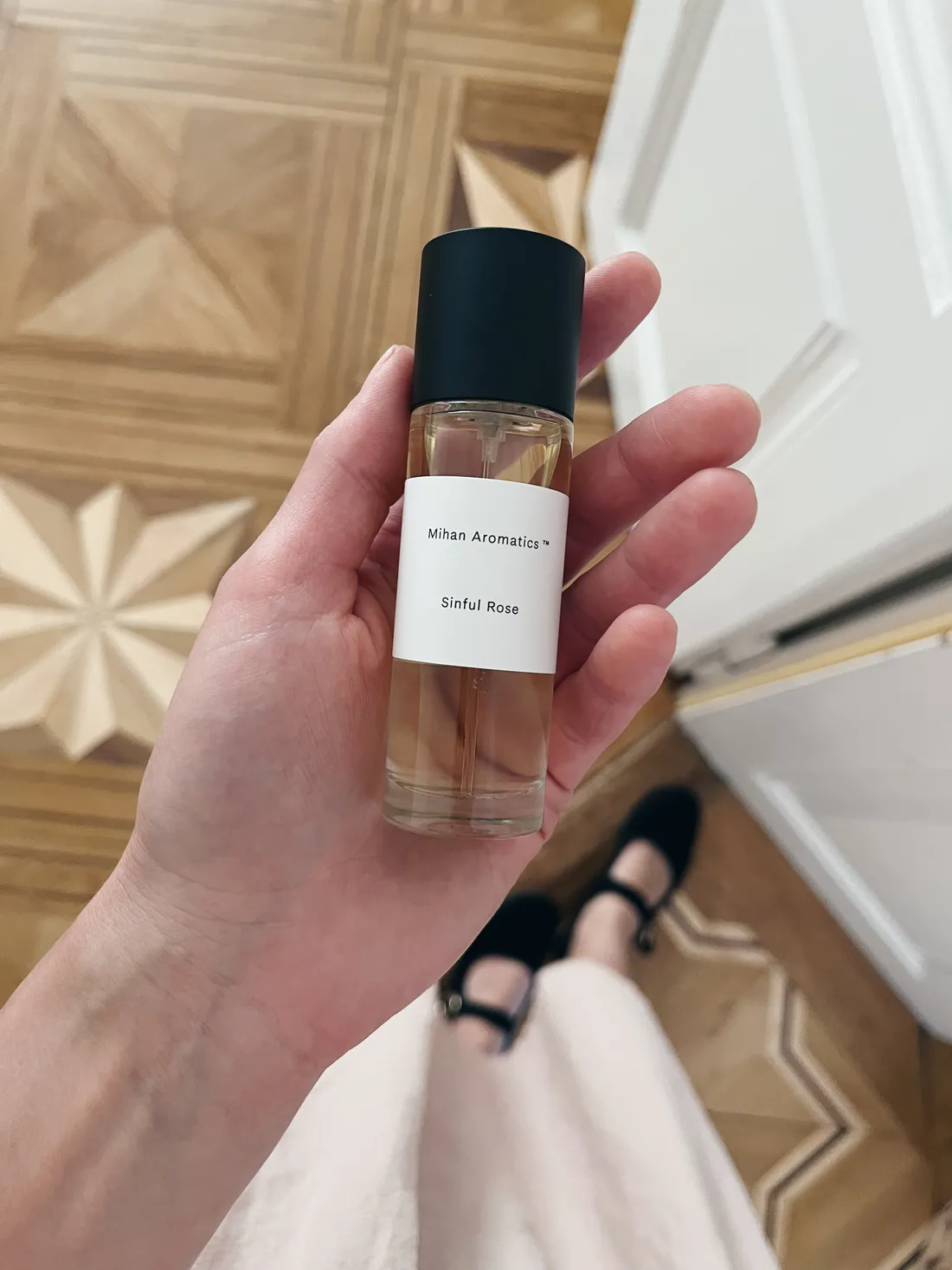 Ruka drží flakón parfému Mihan Aromatics Sinful Rose nad parketami s hvězdicovým vzorem.