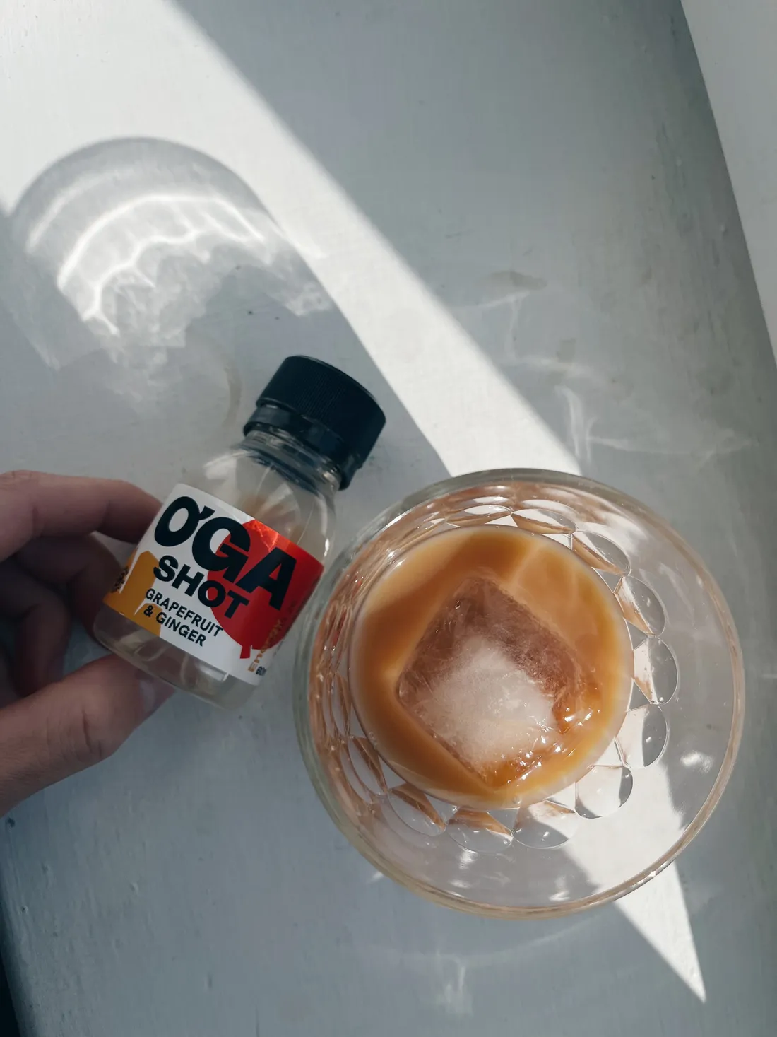 Ruka drží lahvičku O'GA SHOT a sklenici s mléčnou kávou a ledem na světlém stole.