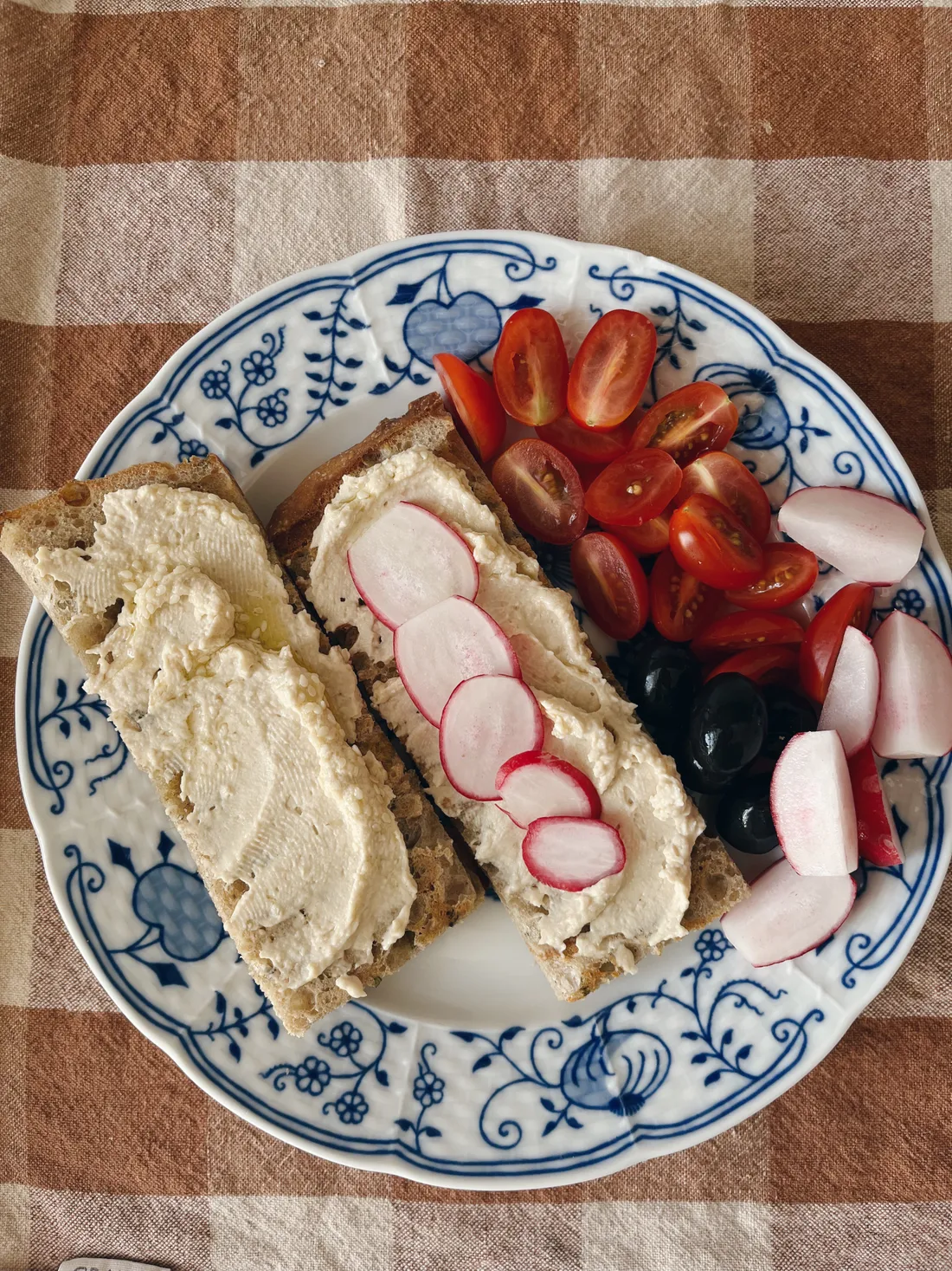 Modrý talíř s dvěma plátky chleba, bílým pomazánkou, ředkvičkami, rajčaty a olivami na kostkovaném ubrusu.