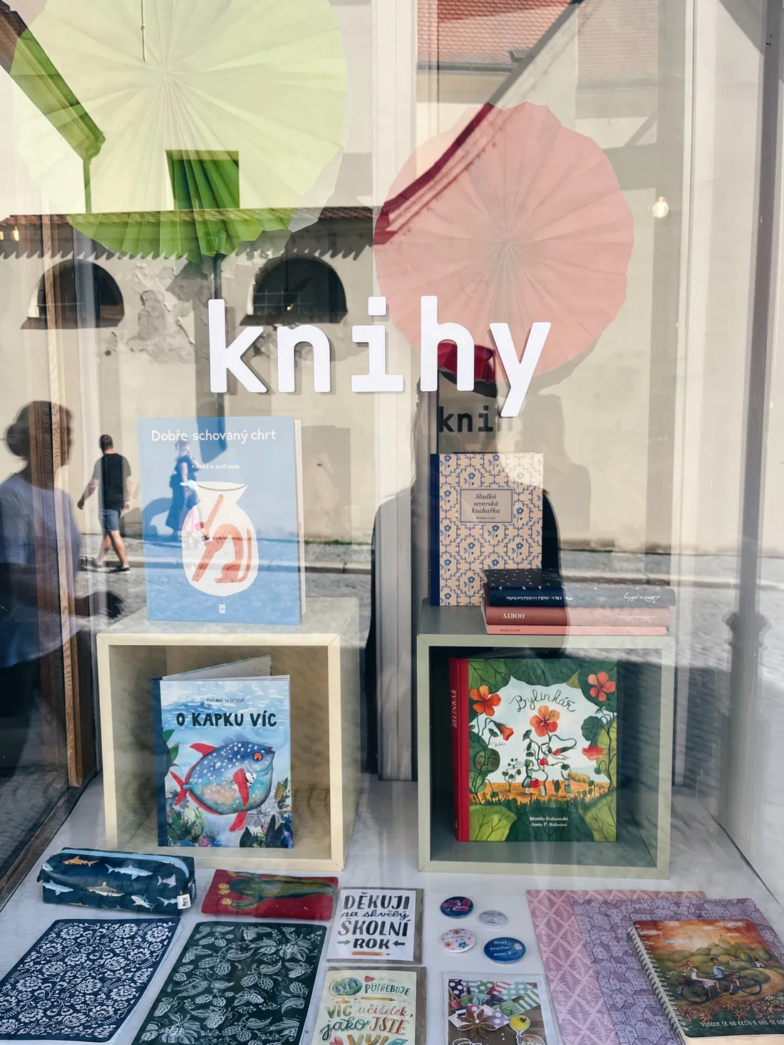 Výloha s nápisem knihy, knihami v boxech, sešity a papírenským zbožím, velké papírové vějíře v pozadí.