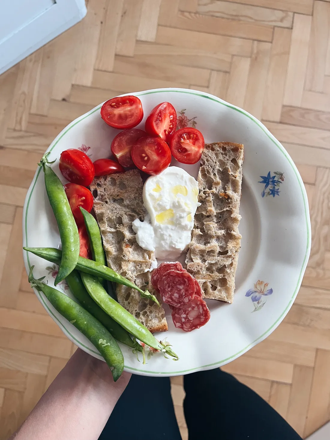 Talíř s toastem, sýrem, cherry rajčaty, hráškem a salámem držený nad parketami shora.