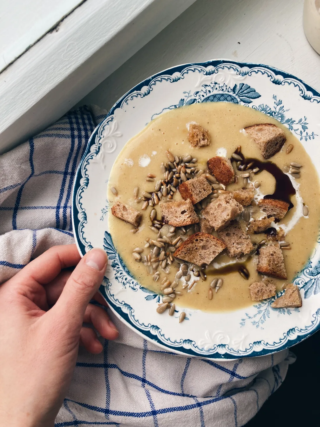 Krémová polévka s krutony, slunečnicovými semínky a olejem na modře zdobeném talíři.