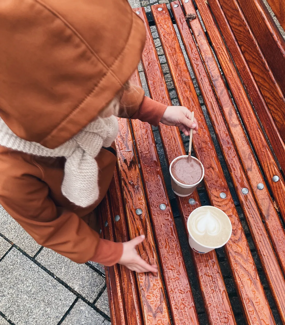 Dítě v teplé bundě míchá horkou čokoládu na mokré dřevěné lavičce, vedle cappuccino.