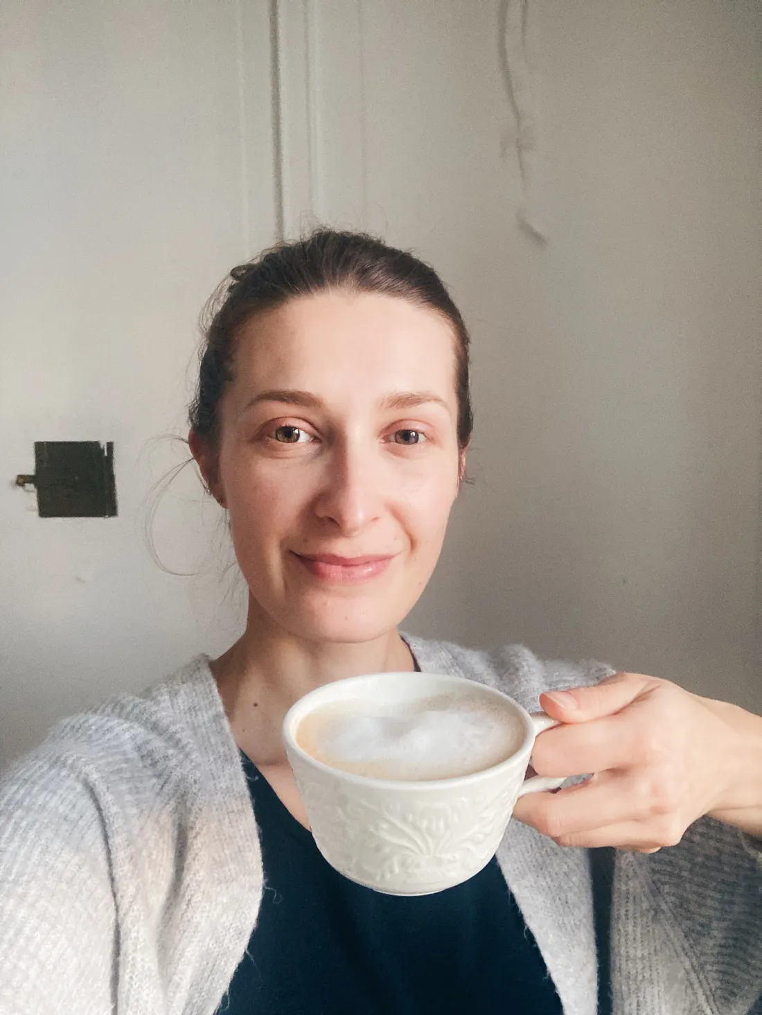 Usmívající se žena drží šálek cappuccina v bílém hrnku s reliéfním vzorem.