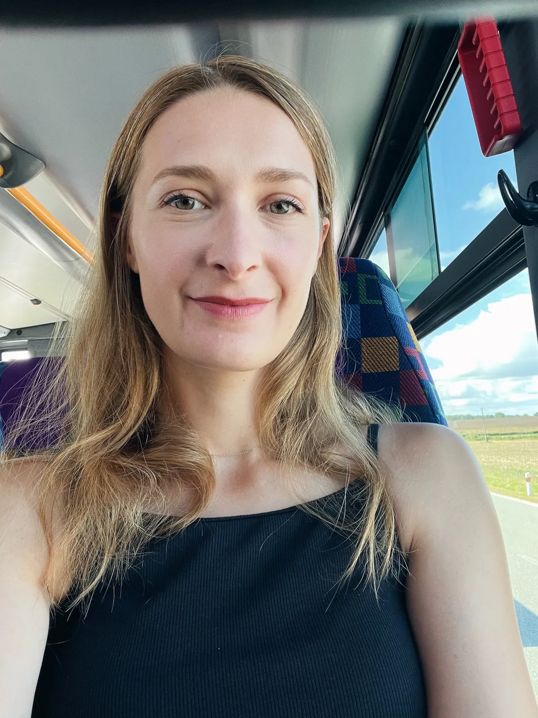 Usmívající se žena v autobuse s kostkovaným sedadlem, venku zelená krajina.