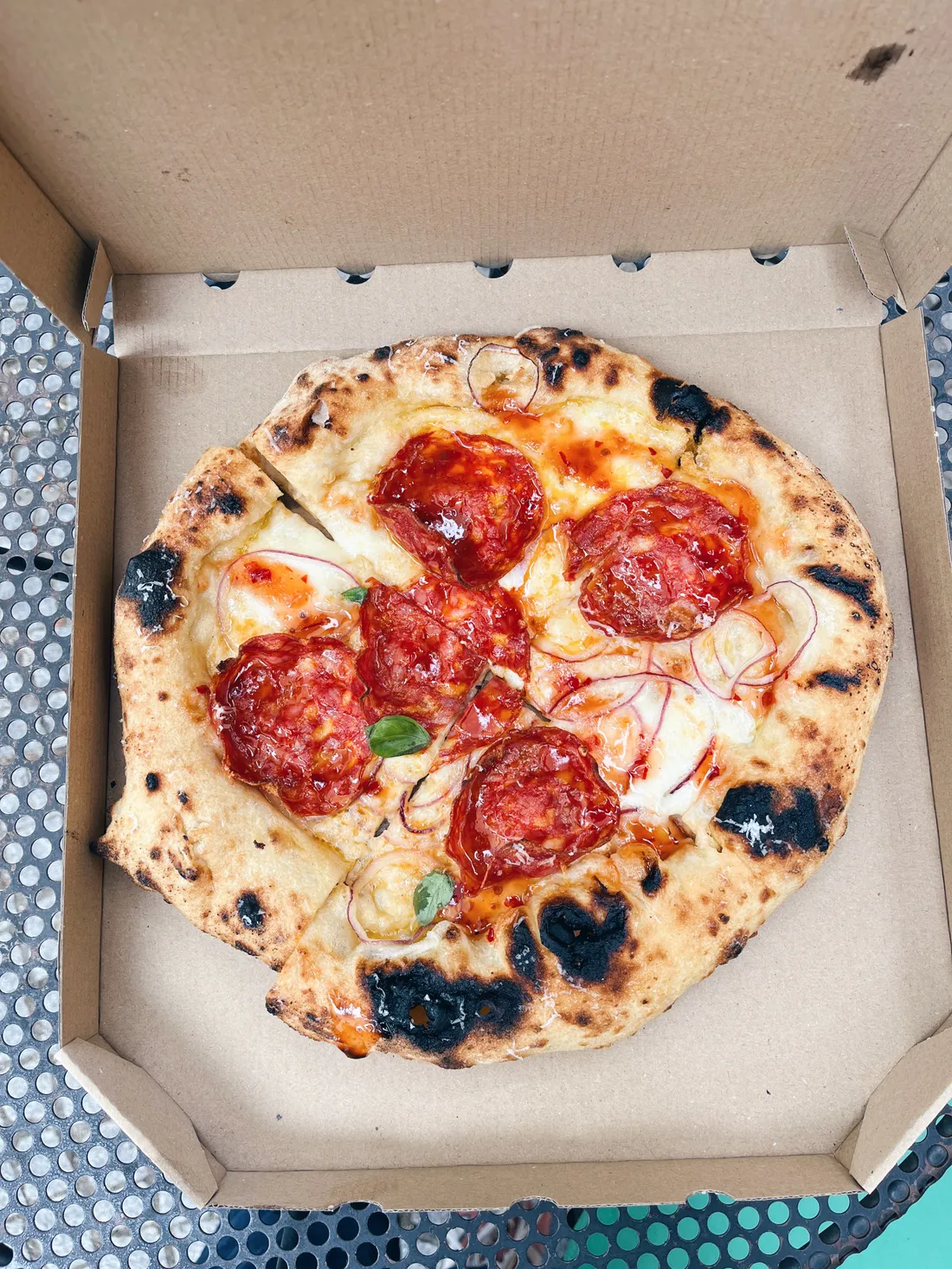 Neapolská pizza se salámem, cibulí, bazalkou a chilli olejem v otevřené papírové krabici na kovovém stolku.