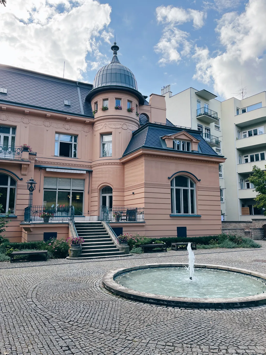 Růžová historická vila s kulatou věží, kašnou a upravenými záhony na dlážděném nádvoří.
