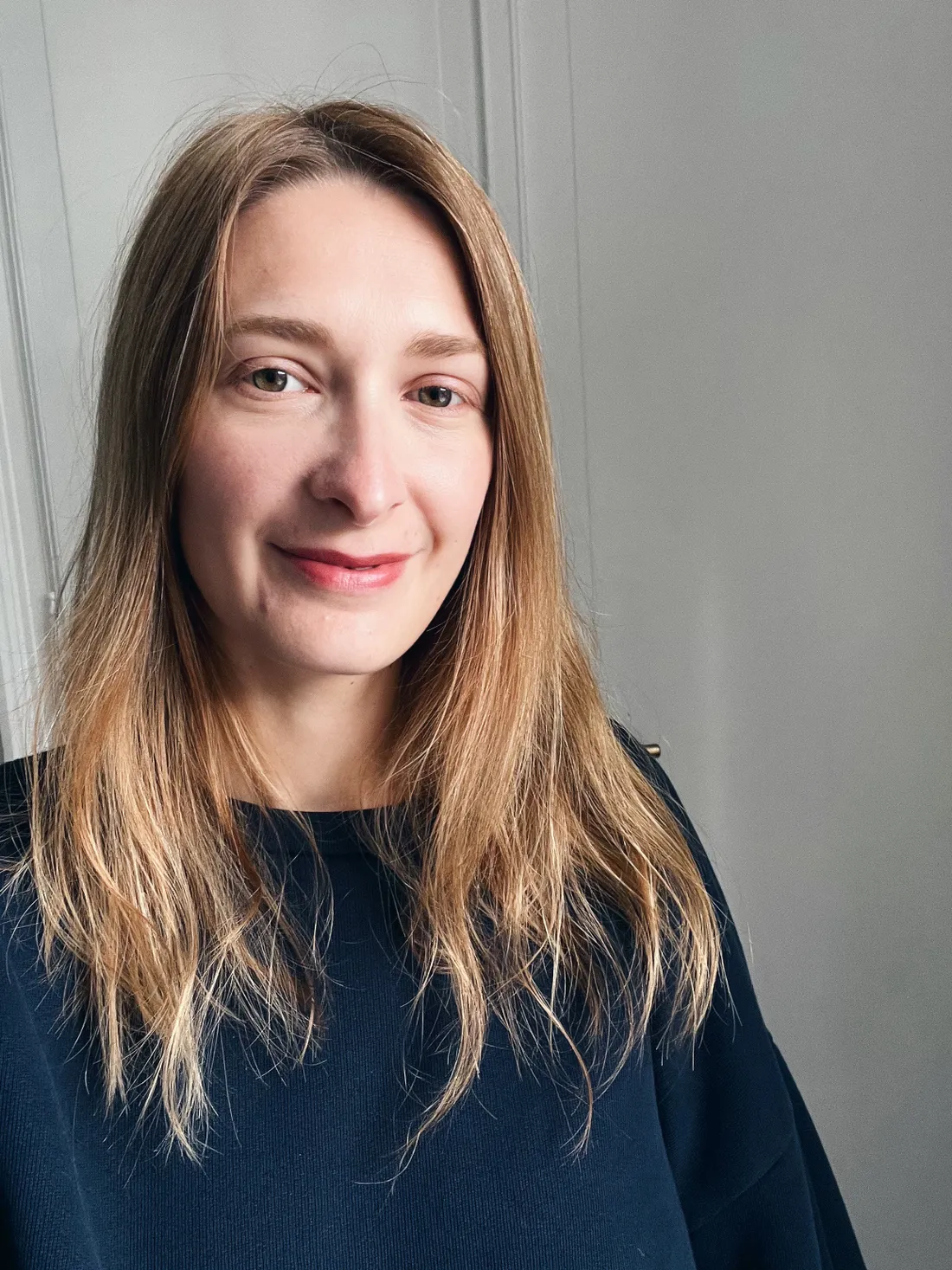 Žena s dlouhými blond vlasy v tmavě modrém triku proti světlé stěně.
