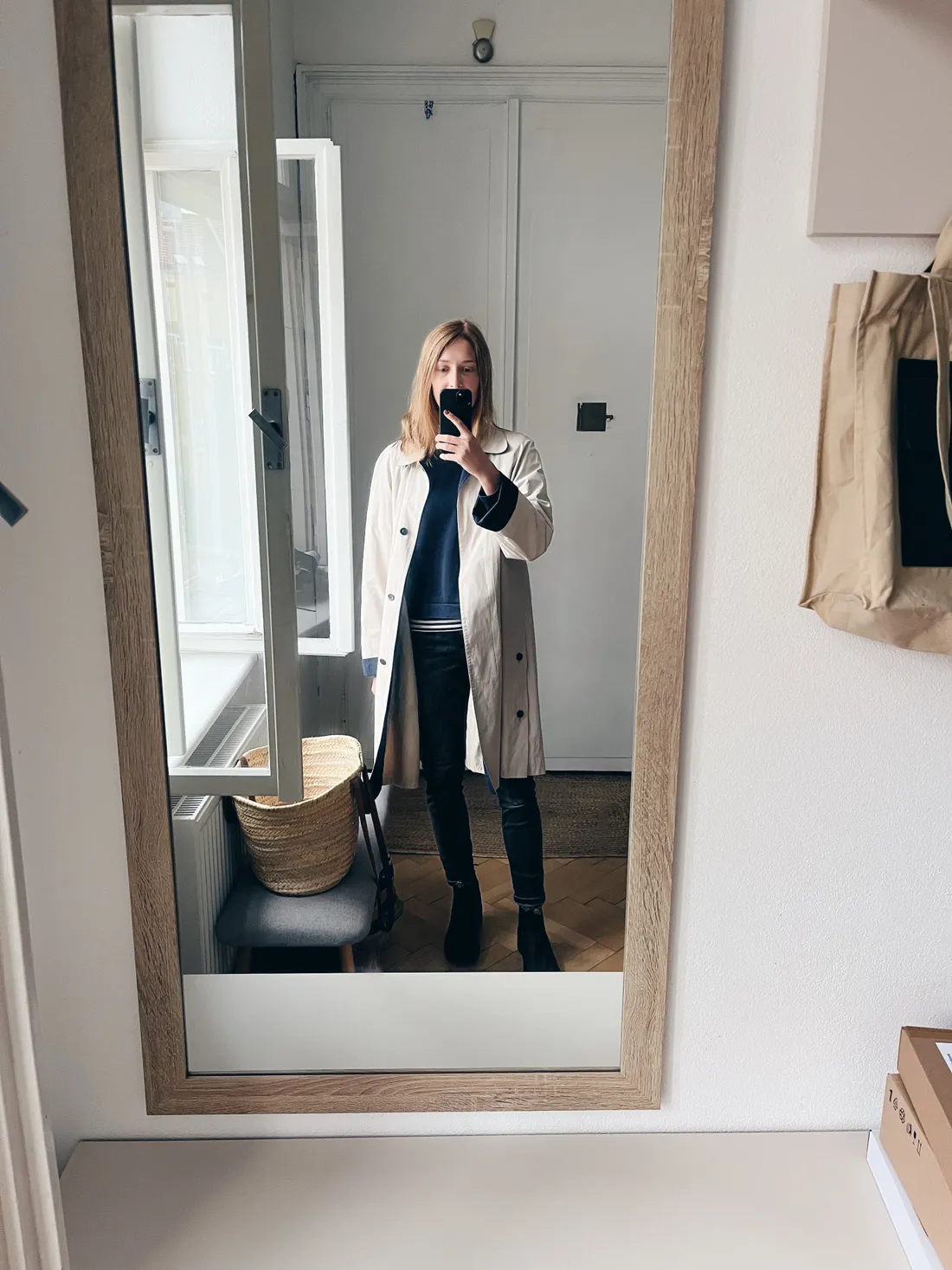 Žena v béžovém trenčkotu a tmavém svetru si fotí selfie v zrcadle se světlým interiérem.