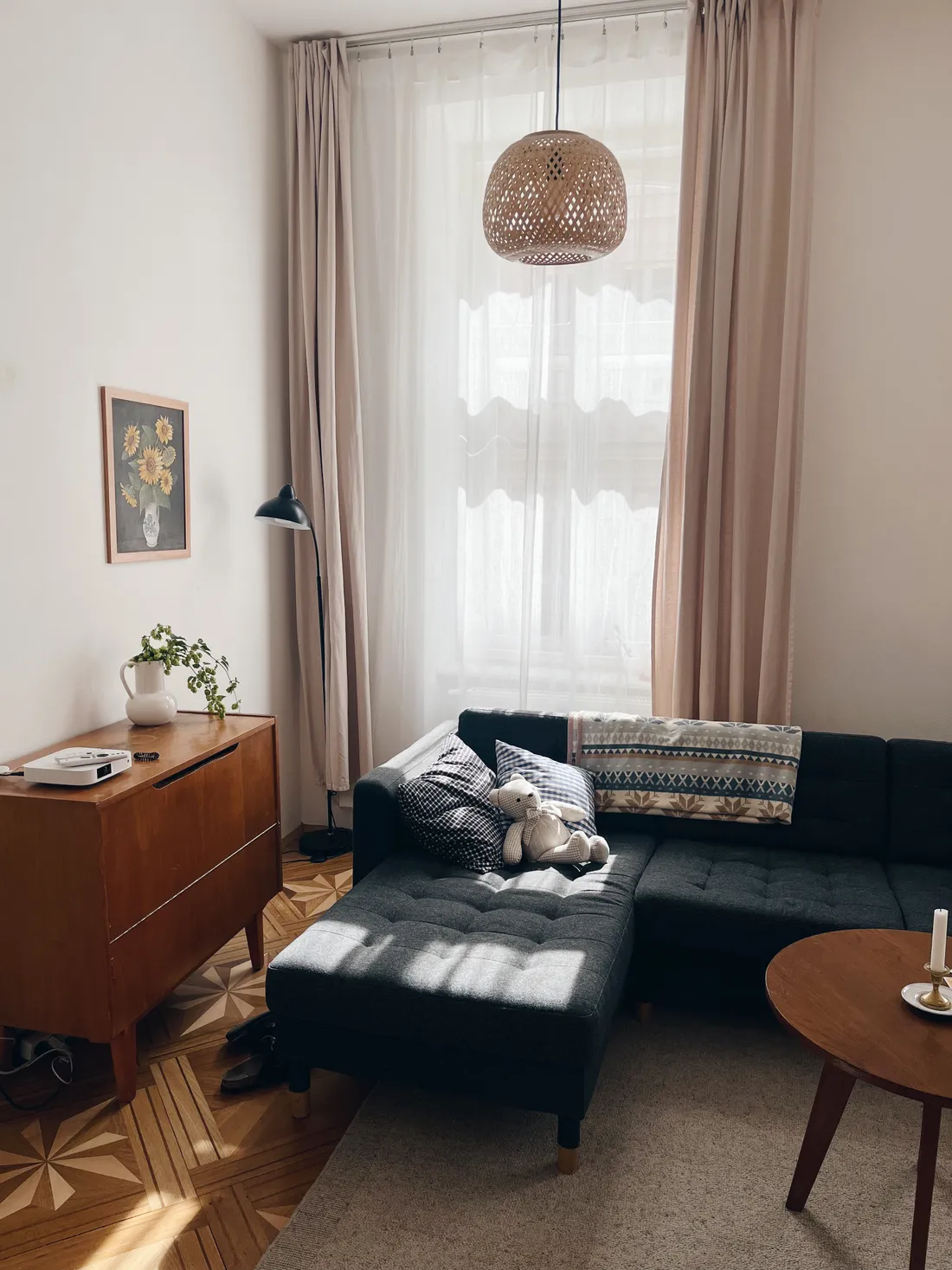 Obývací pokoj se šedou sedačkou, kyticí slunečnic, prosluněným oknem a parketami s hvězdicovým vzorem.