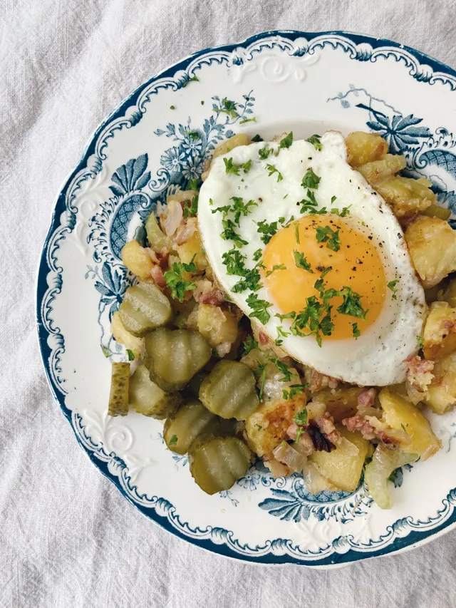 Pytt i panna s restovanými bramborami, slaninou, volským okem a nakládanými okurky na talíři.