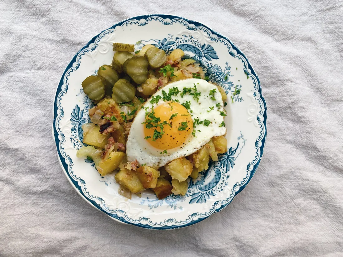Švédská pytt i panna s bramborami, masem a sázeným vejcem s petrželkou, okurky na modrém talíři.