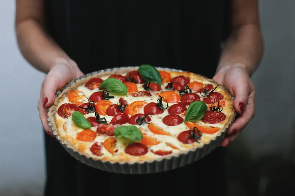Domácí rajčatový quiche s cherry rajčaty a bazalkou v křehkém korpusu, držený v rukou s červenými nehty.