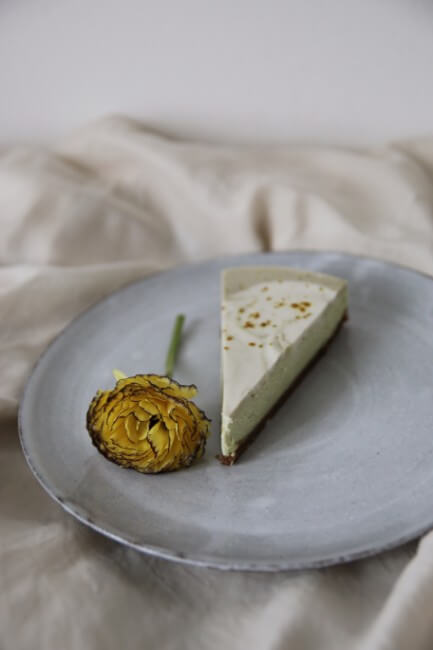 Řez raw avokádového cheesecaku se světle zelenou vrstvou a žlutým květem na keramickém talíři.