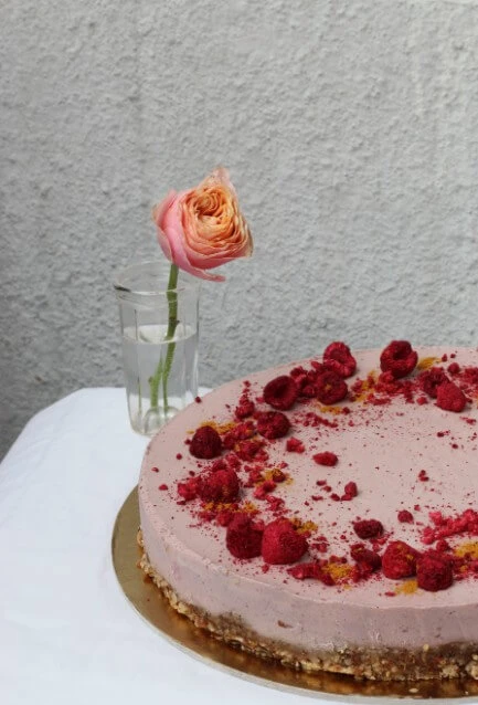 Raw malinový cheesecake s lyofilizovanými malinami na zlatém podkladu, růže ve sklenici v pozadí.