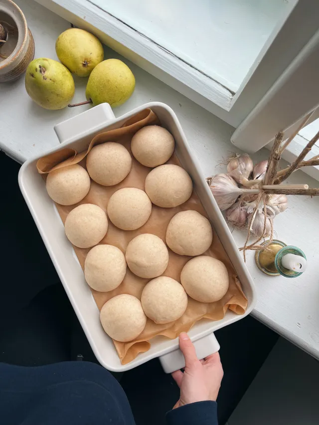 Dvanáct kynutých bochánků v bílé kameninové formě na papíře, hrušky, česnek a svíčka na okenním parapetu.