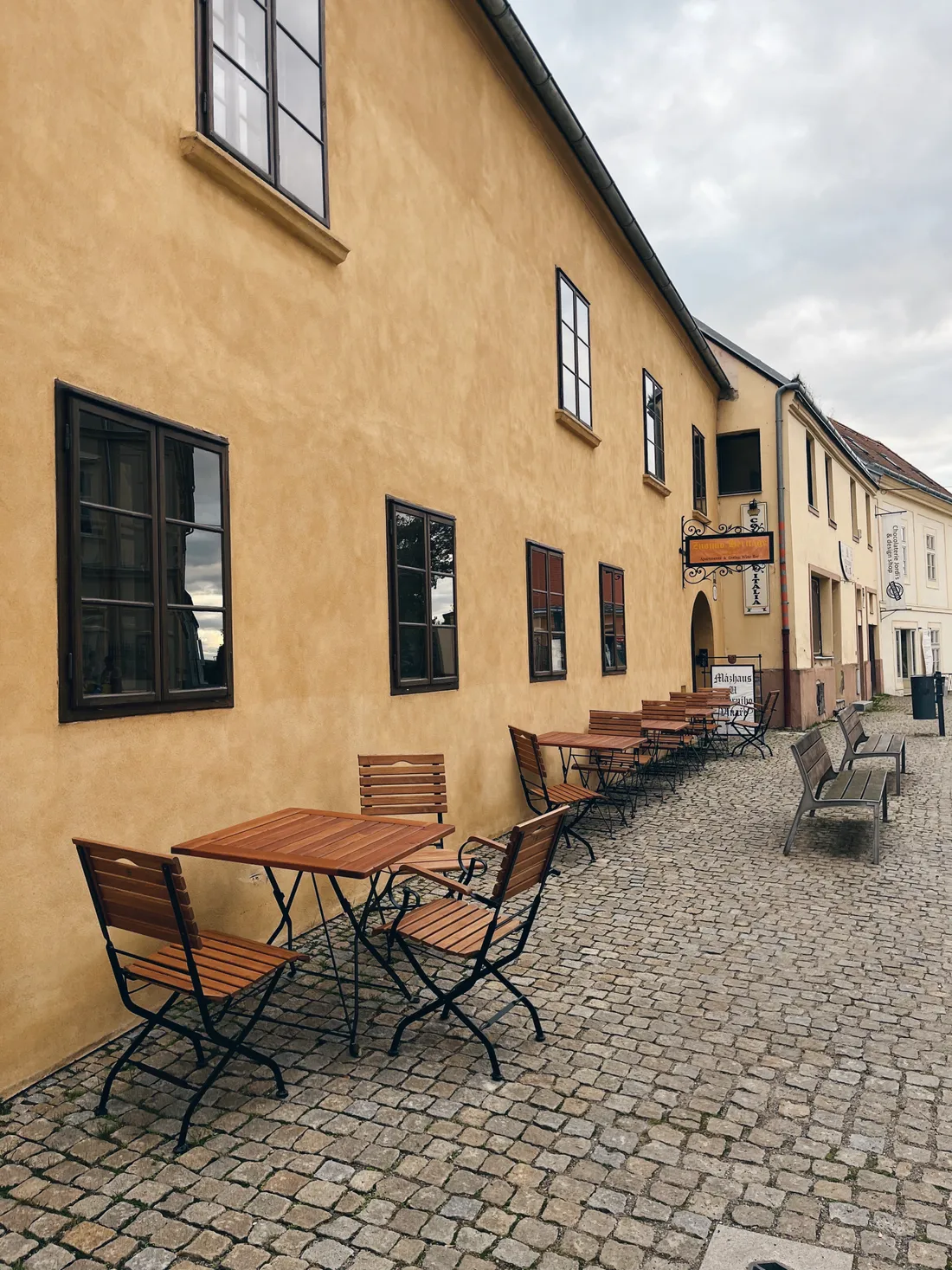 Venkovní posezení před dlouhou žlutou historickou budovou na dlážděné ulici