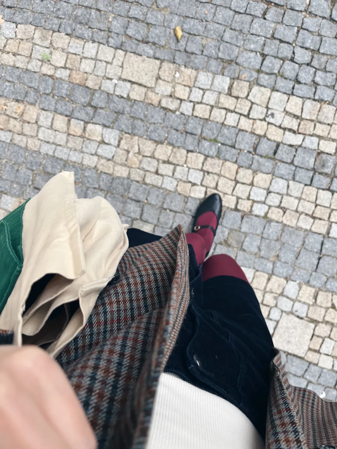 Pohled shora na kárované sako, manšestrovou sukni a vínové punčochy na dláždění