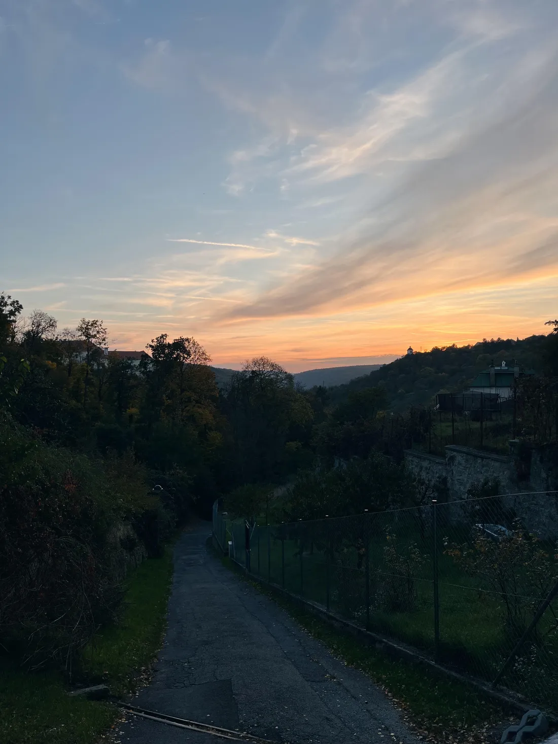 Večerní pohled na údolí při západu slunce, cesta mezi zahradami a silueta rotundy na kopci
