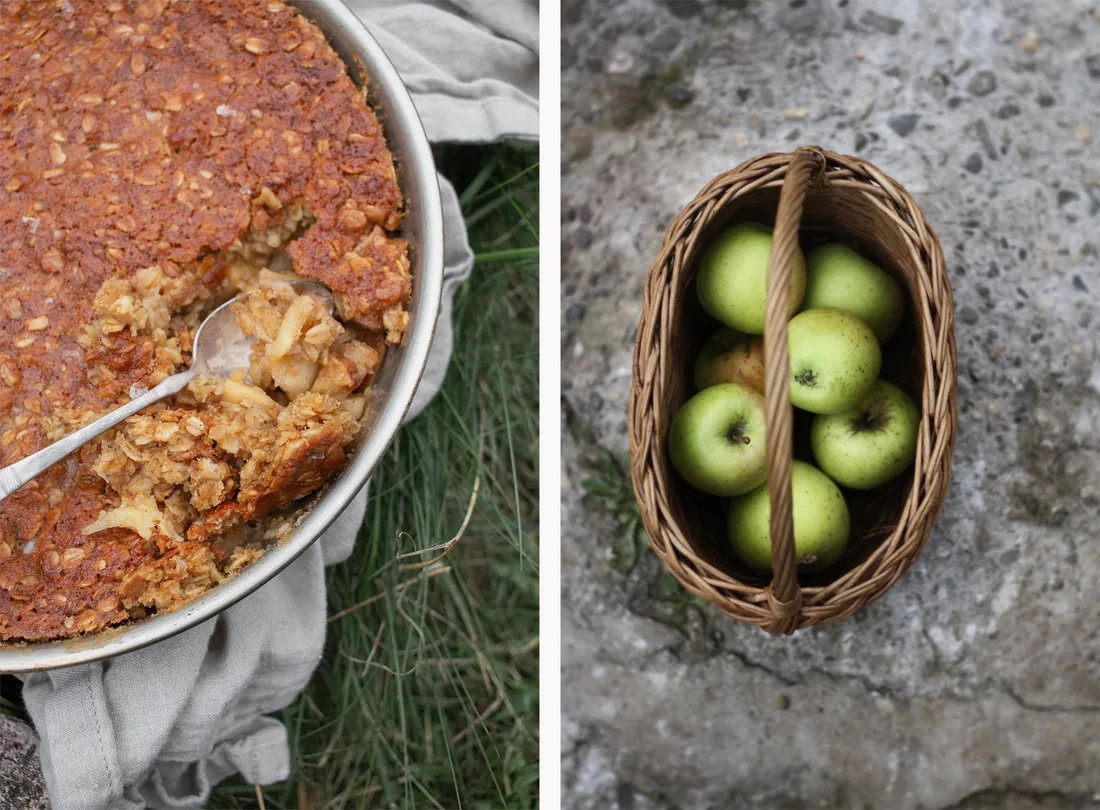 Jablečný crumble v plechu se lžící a proutěný košík se zelenými jablky na trávě a kameni.