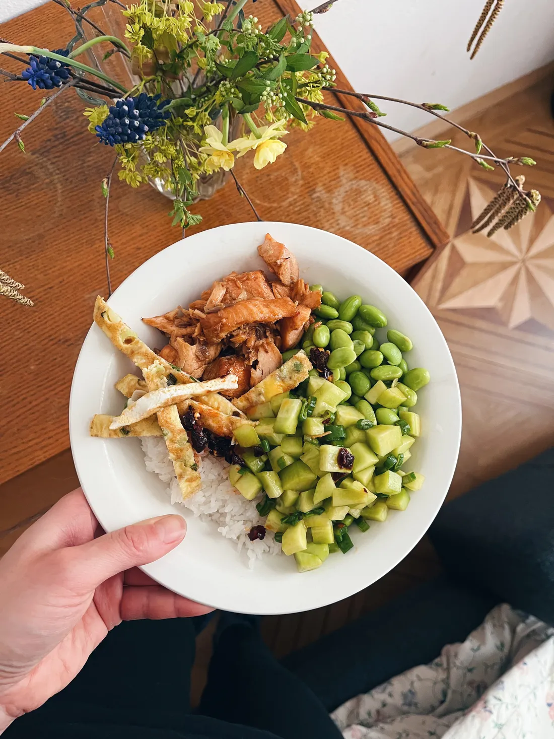Miska rýže s lososem, edamame, okurkou a proužky omelety držená nad dřevěným stolem s jarní kyticí.