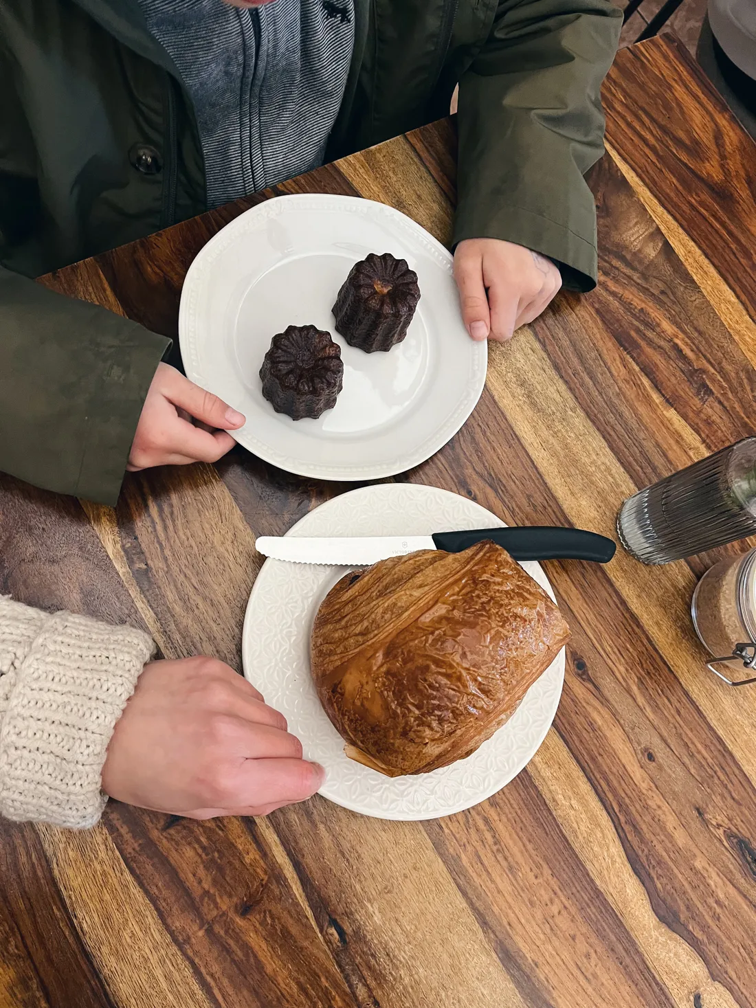 Dva talíře pečiva na dřevěném stole — cannelés a velký máslový závin, ruka drží talíř.