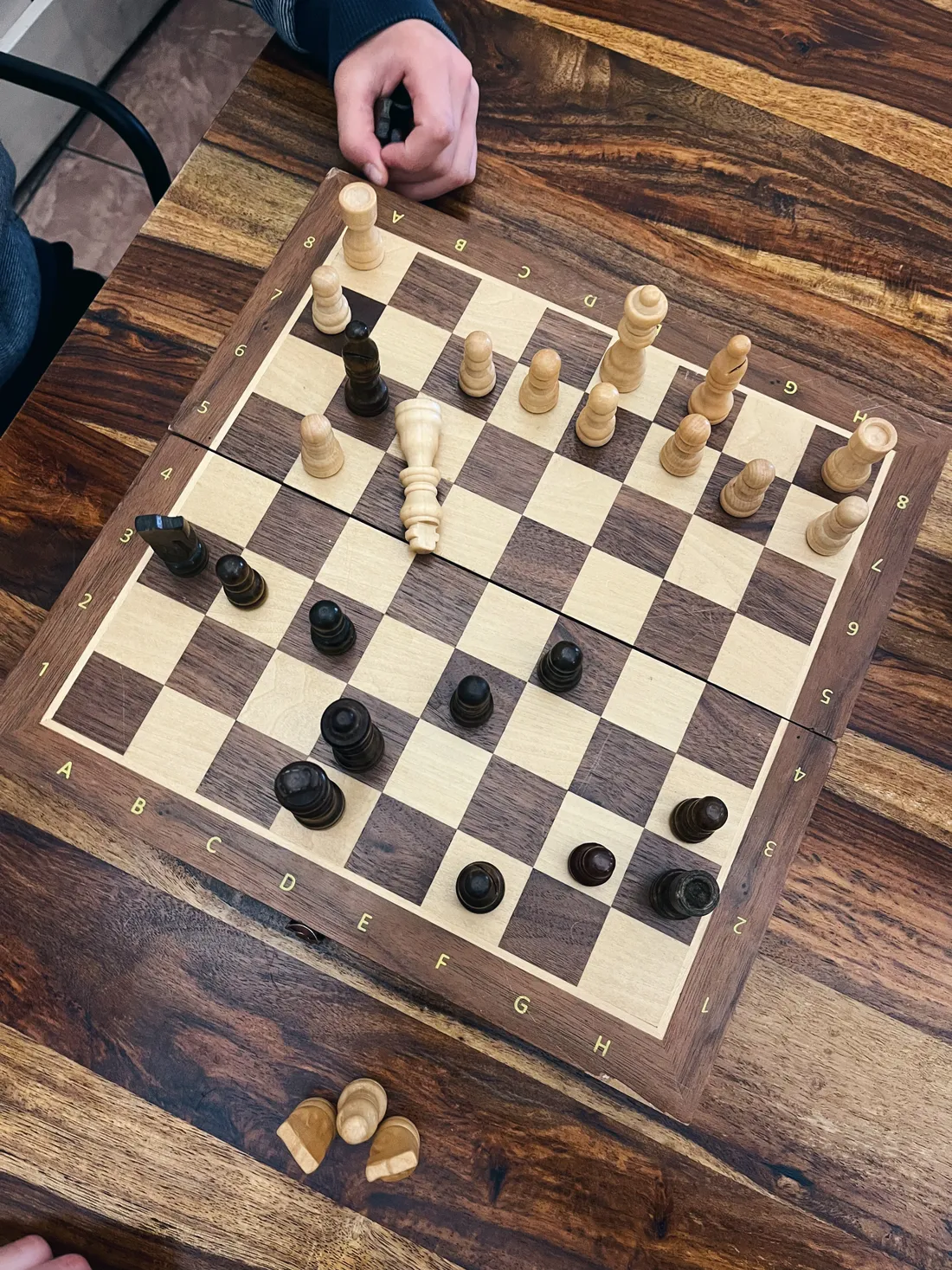 Dřevěná šachovnice s rozestavenými figurkami, ruka hráče a vyřazené figurky na stole.
