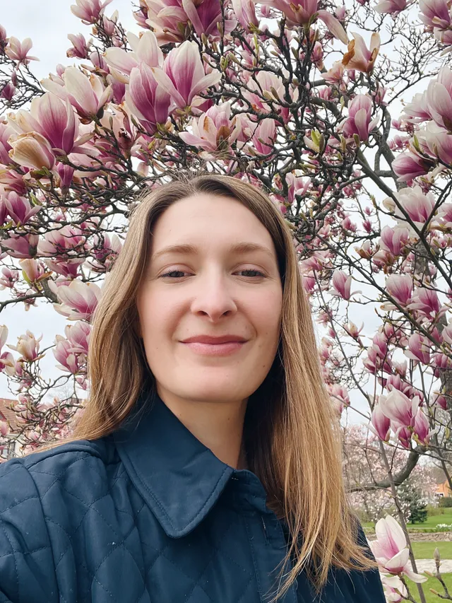Usmívající se žena v modré prošívané bundě před rozkvetlou magnolií s růžovými květy.