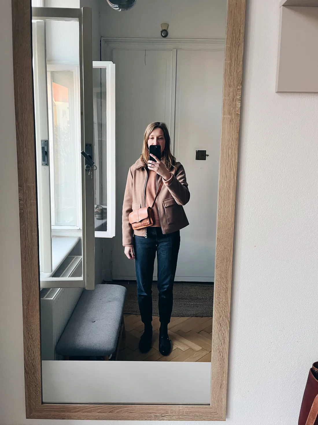 Selfie v zrcadle — žena v hnědé bundě s kožíškem, tmavých džínách a kožené crossbody kabelce.