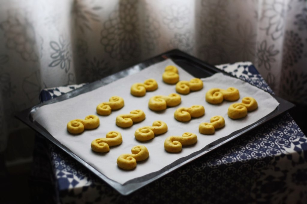 Syrové šafránové lussekatter v řadách na plechu s pečicím papírem na modrém ubrusu.