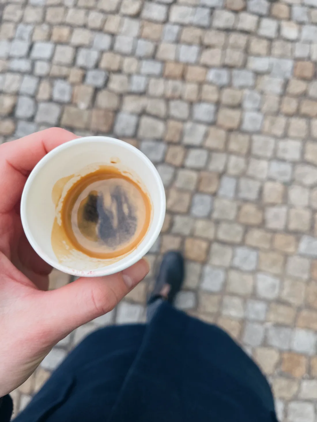 Ruka drží malý papírový kelímek espresso nad šedým dlážděným chodníkem, noha v černé botě.