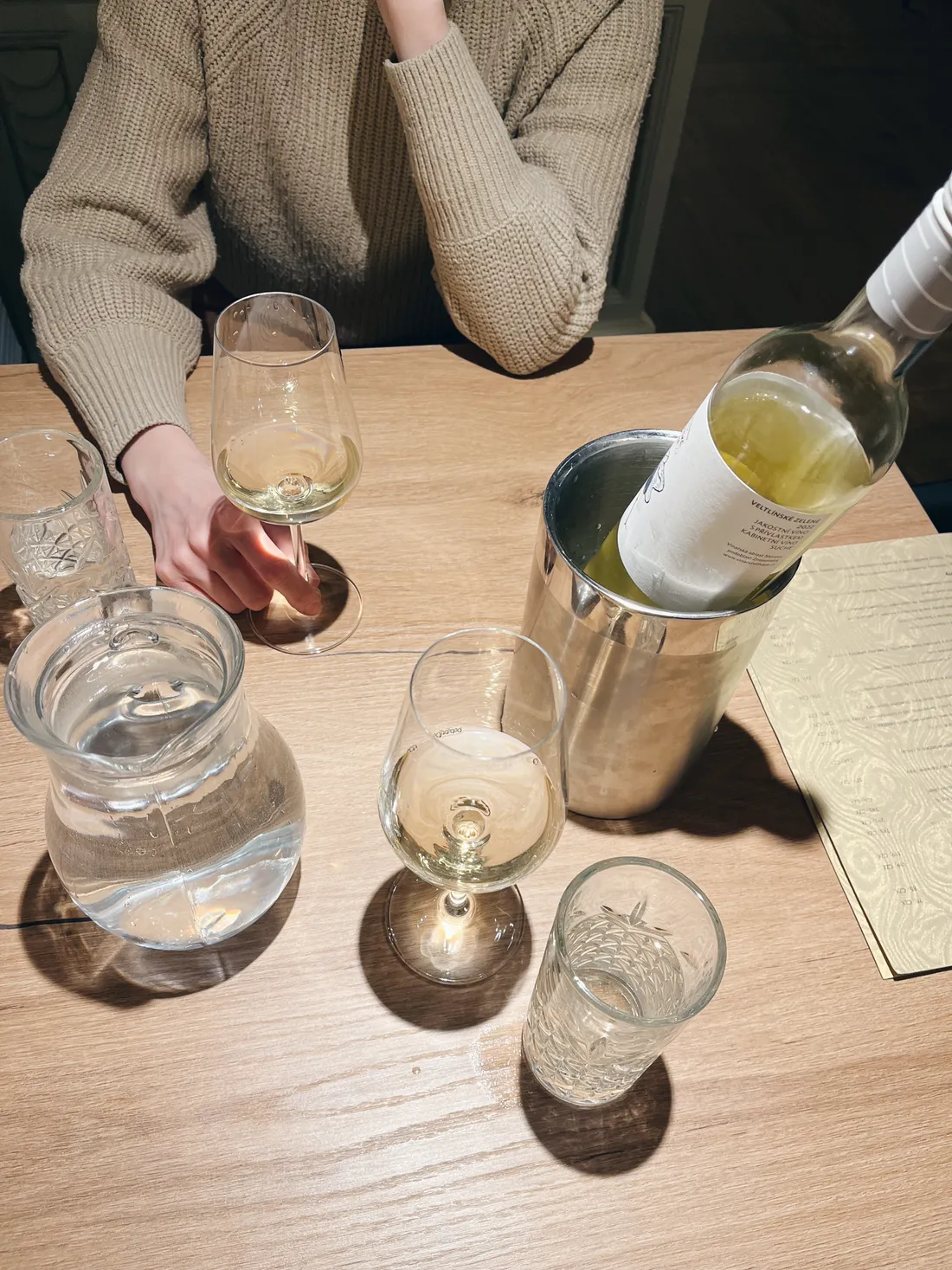 Dva sklenice bílého vína a láhev v chladiči na dřevěném stole s karafou vody a menu.