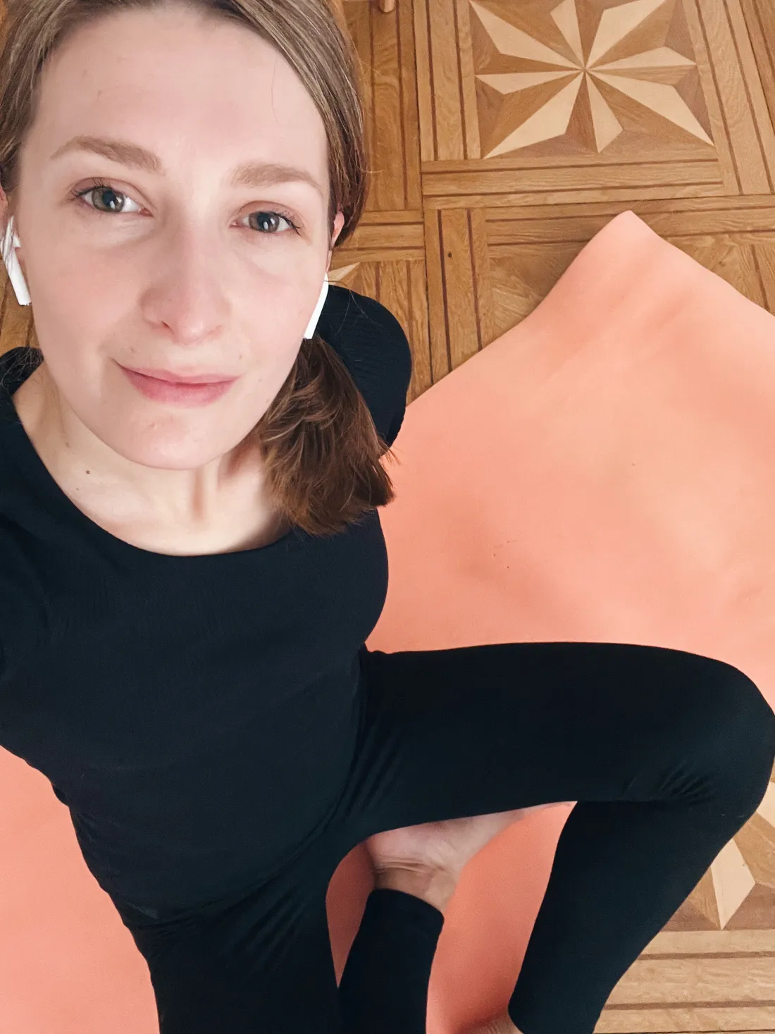 Žena s bílými sluchátky sedí na růžové podložce na parketách s hvězdičkovým vzorem.