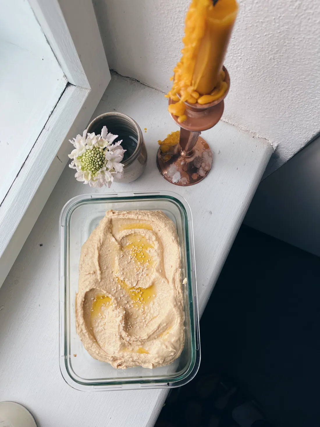 Hummus s olivovým olejem a sezamem v skleněné nádobě na parapetu se svíčkou a květinou.