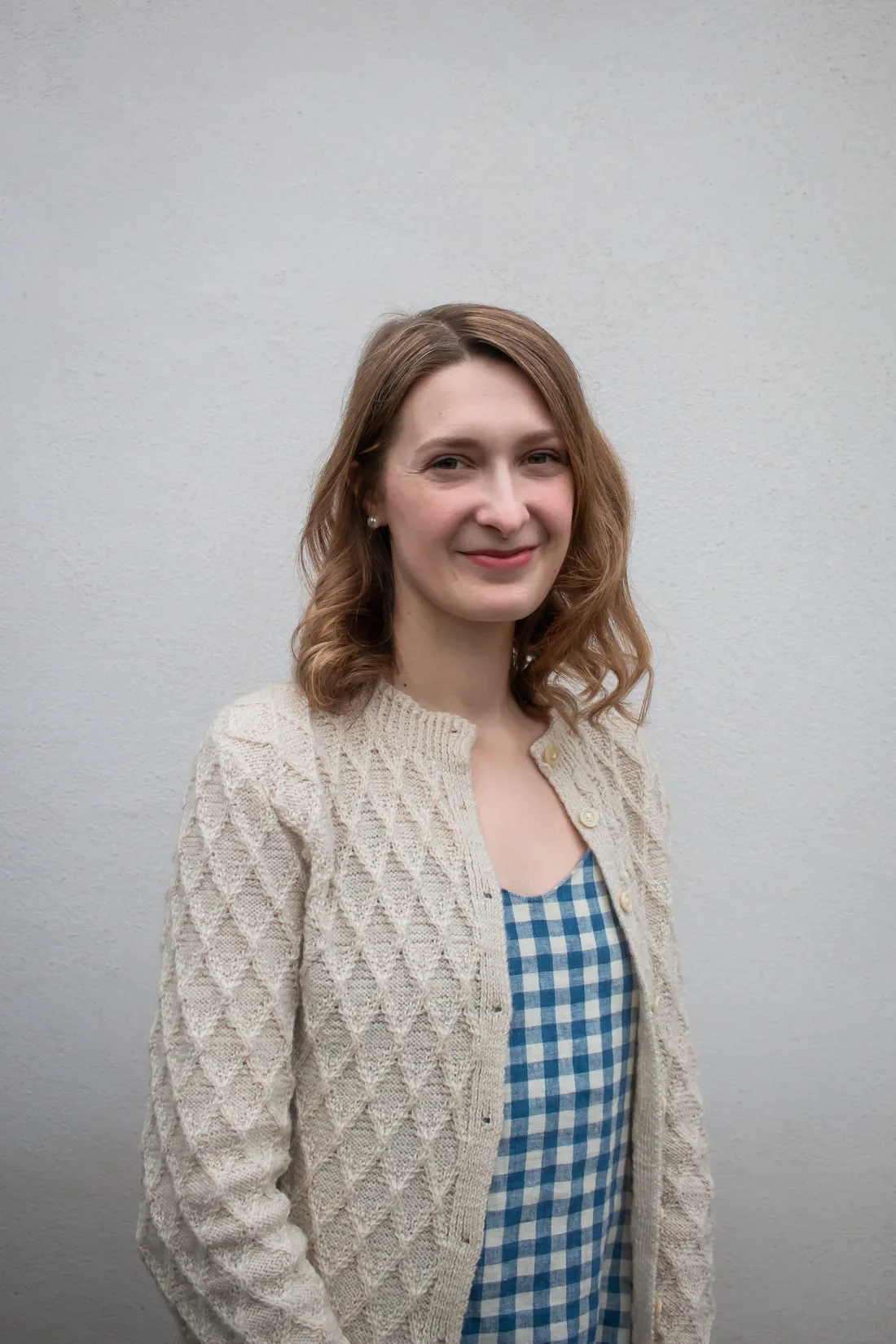 Blondýna v krémovém kardiganu s kosočtvercovým vzorem a modro-bílé kostkované halence u šedé stěny.
