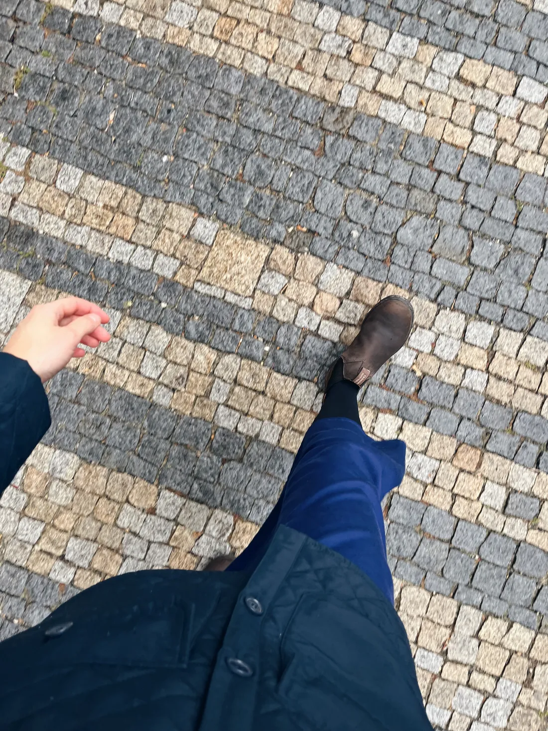 Nohy v modrých kalhotách a hnědých botách kráčejí po dlážděném chodníku s pruhy kostek.
